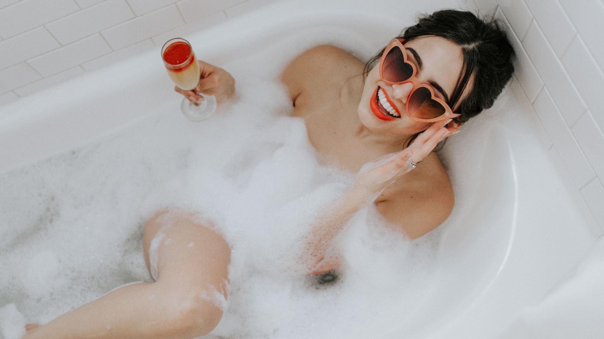 A Woman In A Bathtub With A Drink And A Man In A Mask